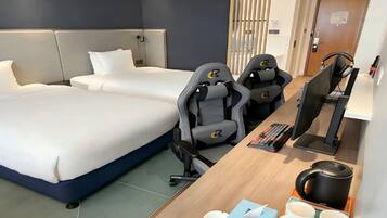 A hotel room with two beds, a desk with a computer, and gaming chairs.