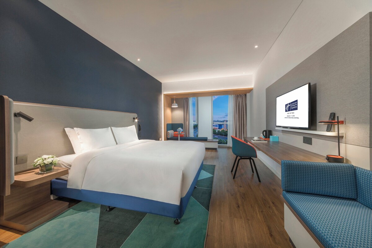 A modern hotel room with a large bed, a desk, a seating area, and a television.
