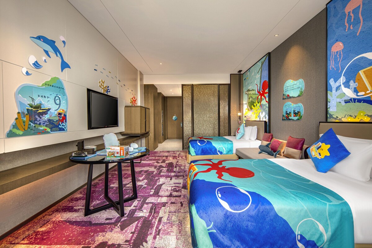 Quarto de hotel com duas camas, televisão, uma mesinha e obras de arte nas paredes.
