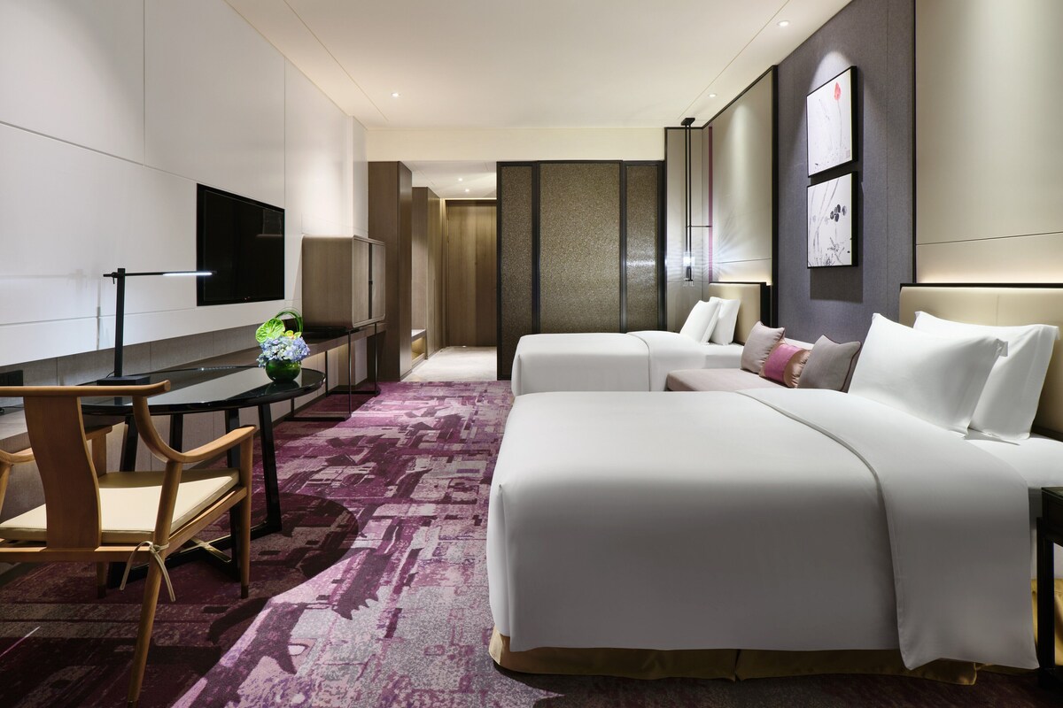 Quarto de hotel com uma cama grande, escrivaninha com cadeira, televisão e um banheiro ao fundo.