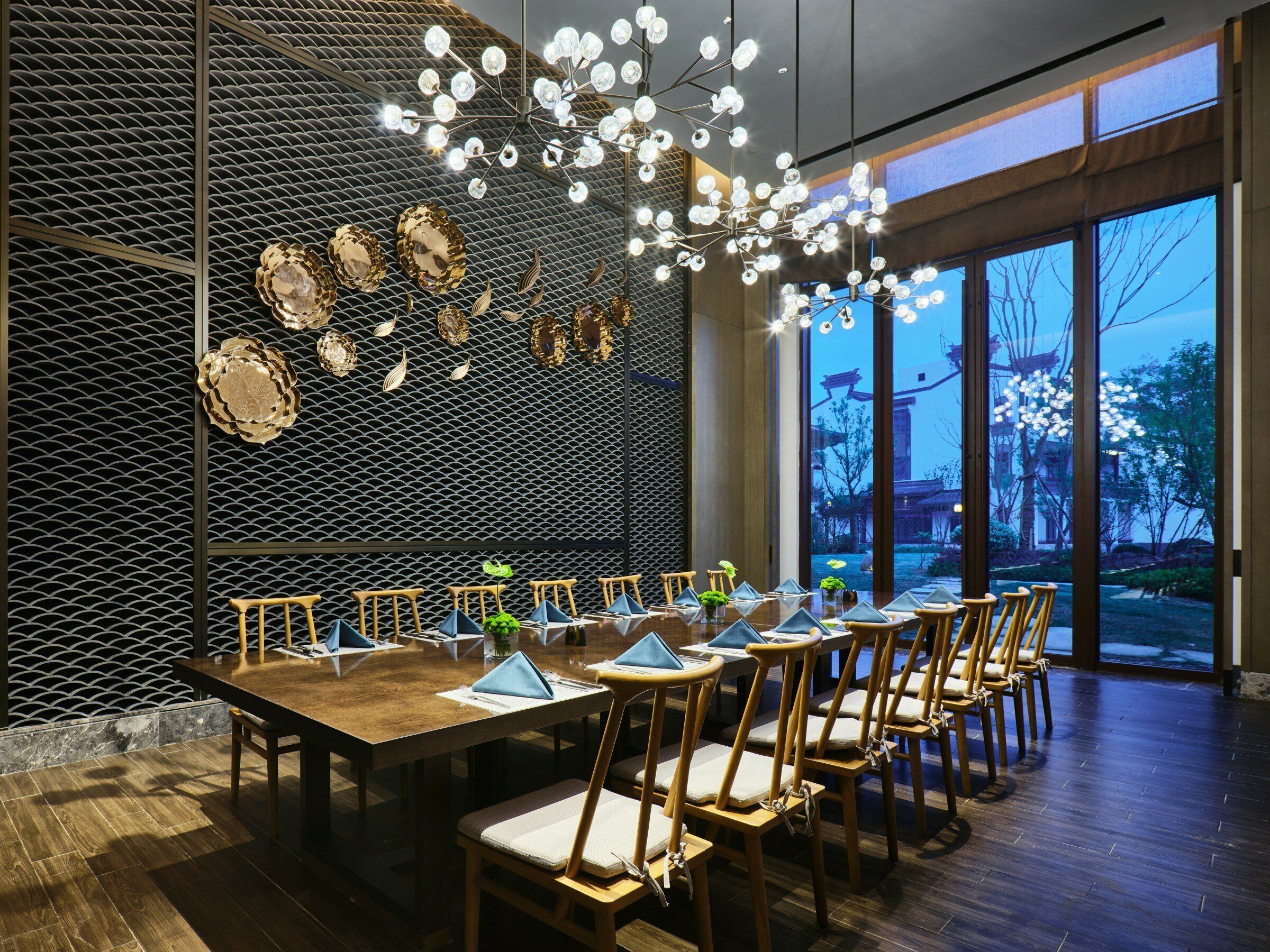 Een eetruimte met een lange tafel, stoelen en een decoratieve wand met hangende lampen.