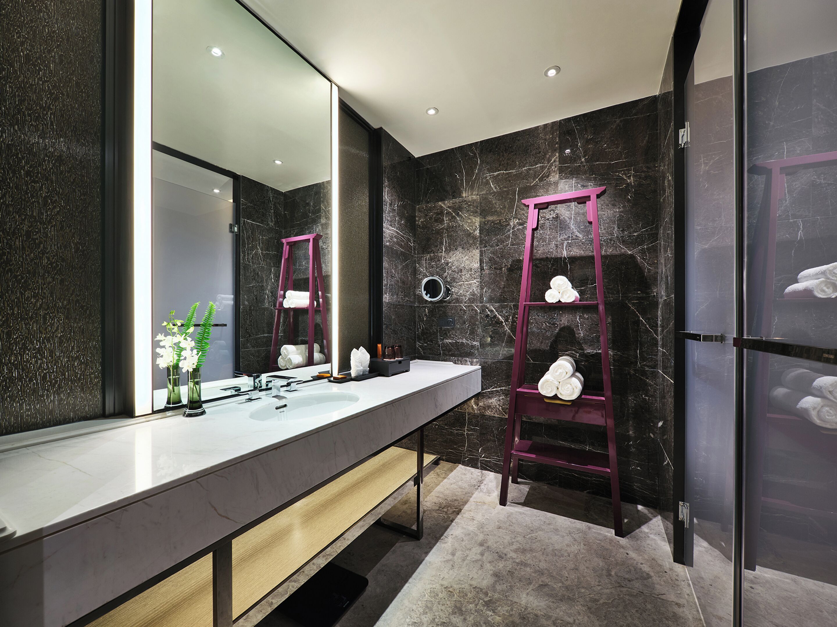 A modern bathroom with a marble vanity, a large mirror, a pink ladder shelf, and a marble wall.