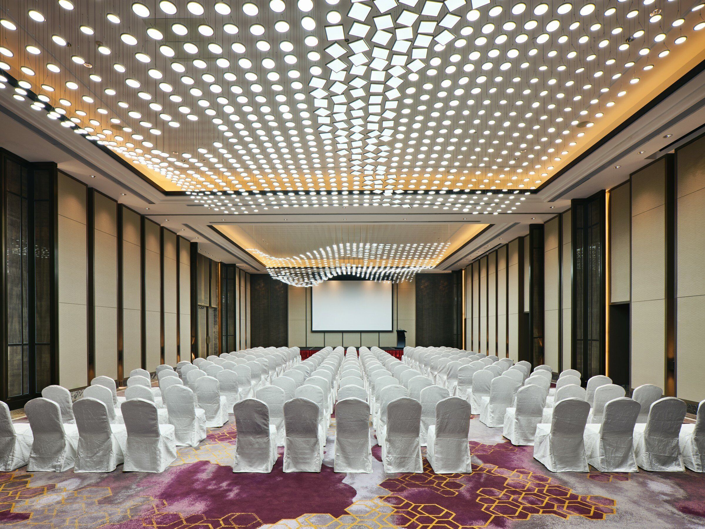 A conference room with rows of chairs, a large screen, and a patterned carpet.
