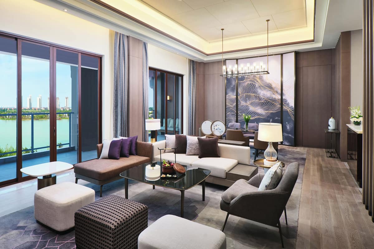 Ein modernes Hotelzimmer mit einem großen Fenster und Blick auf die Stadt, einer gemütlichen Sitzecke mit Sofas und Poufs sowie einem dekorativen Wandbild.