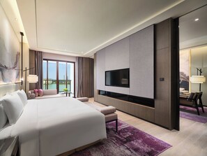 Quarto de hotel moderno com uma cama grande, televisão de tela plana na parede, área de estar com sofá e poltronas, e varanda com vista para a cidade.