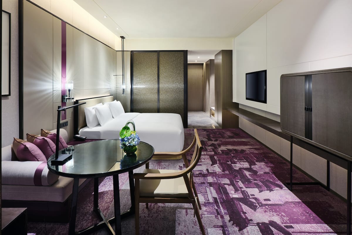 Ein modernes Hotelzimmer mit Bett, einer Sitzecke mit Glastisch, einem Fernseher und einem großen Fenster.
