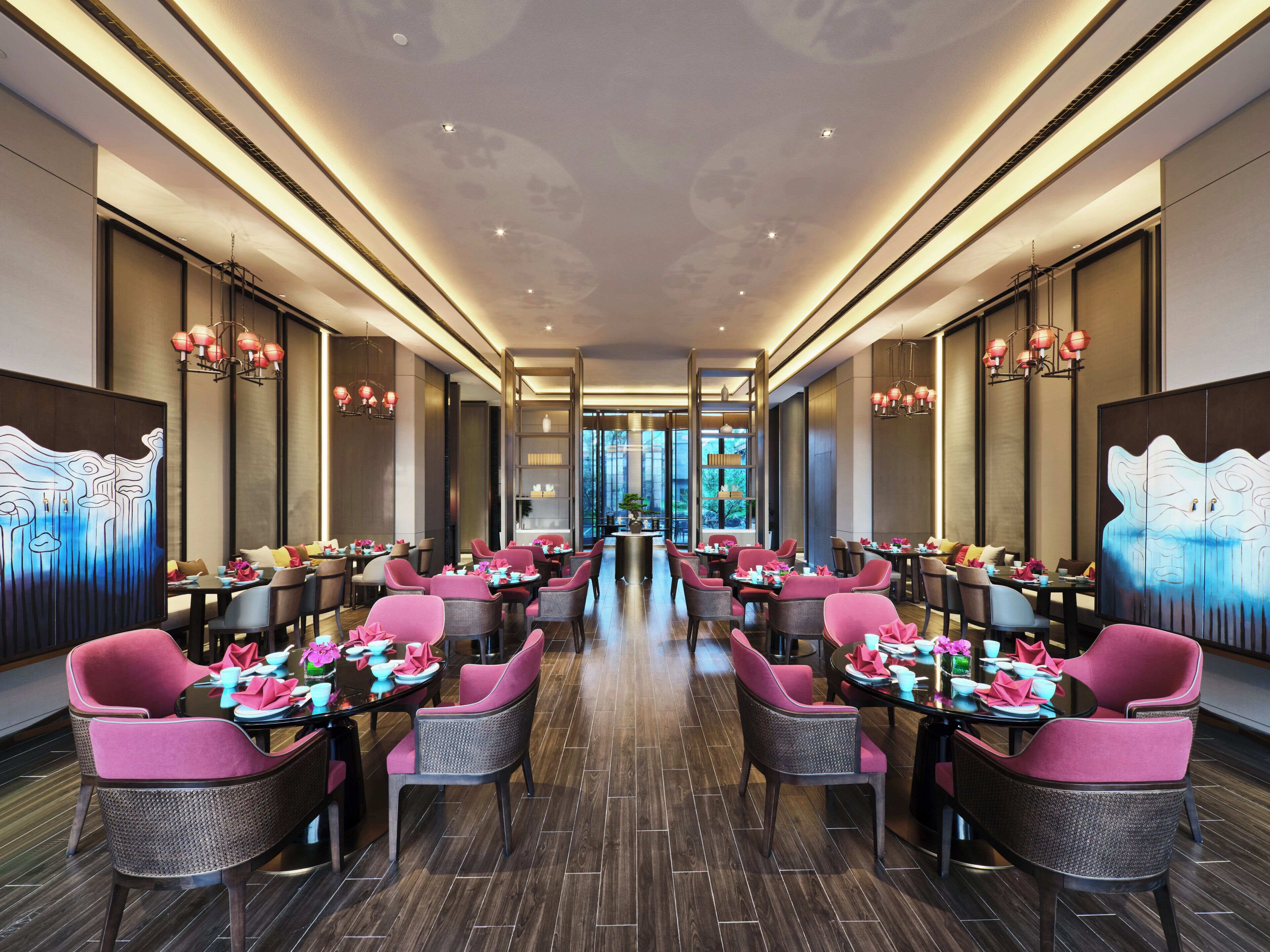 Ein modernes Restaurant mit einer zentralen Gäng, Tischen mit rosafarbenen Stühlen und dekorativer Beleuchtung.