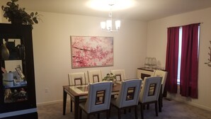 A dining area with a glass-top table, six chairs, a chandelier, a floral painting, and a window with red curtains. - Spacious 5 Bedroom rental (Waukesha)