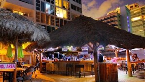 An outdoor bar area with thatched umbrellas, a pool, and a view of a modern building in the background. - Oceanfront Studio w/balcony & Private Beach - Walk to shops & Dining (Fort Lauderdale)