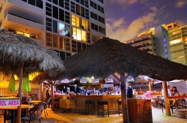 An outdoor bar area with thatched umbrellas, a pool, and a view of a modern building in the background. - Oceanfront Studio w/balcony & Private Beach - Walk to shops & Dining (Fort Lauderdale)