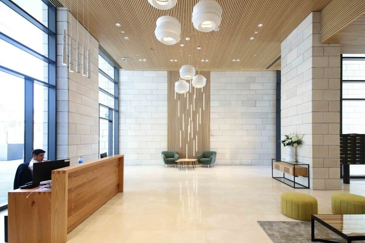 Eine großzügige Lobby mit einem Holzbuchttisch, zwei grünen Sessel und einem modernen Deckenbau mit Pendelleuchten.