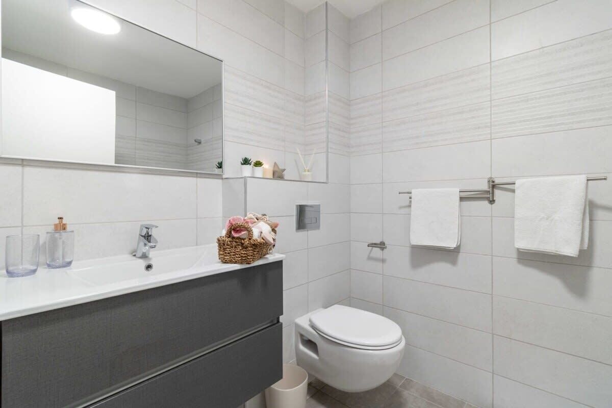 A modern bathroom with a white sink, mirror, and toilet. There are towels hanging on a rack and a basket of toiletries on the counter.