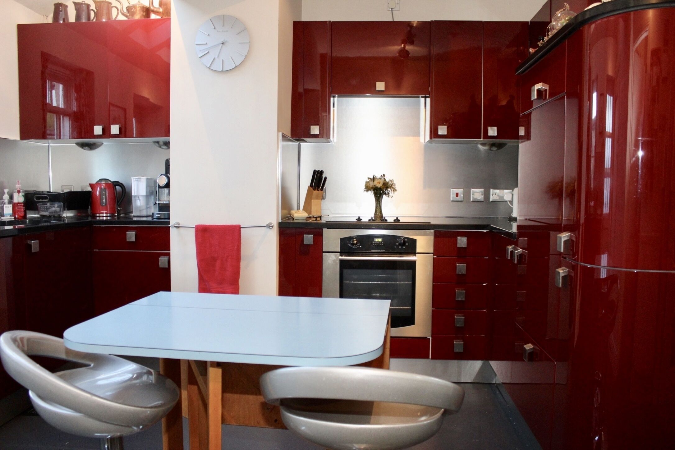 A modern kitchen with red cabinets, a stainless steel refrigerator, a built-in oven, and a white dining table with two chairs.