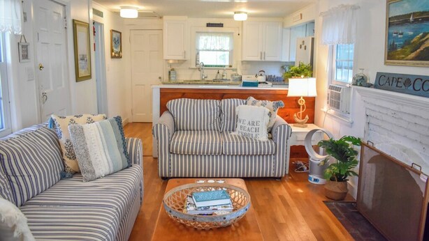 Un salón acogedor con sofás a rayas y una mesa de centro, una cocina con armarios blancos y un ventanal, y una chimenea con un letrero que dice 'Cape Cod'.