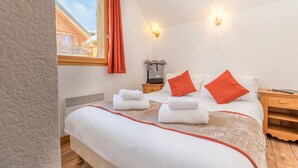 A bedroom with a bed, two bedside tables, a heater, a window with a curtain, and a view of a wooden building. - Appart proche de la navette aux pistes (Les Déserts)