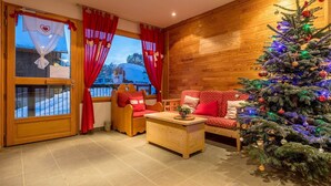 A cozy room with a wooden interior, a wooden sofa with red cushions, a small wooden table with a plant, and a Christmas tree with lights. - Appart proche de la navette aux pistes (Les Déserts)