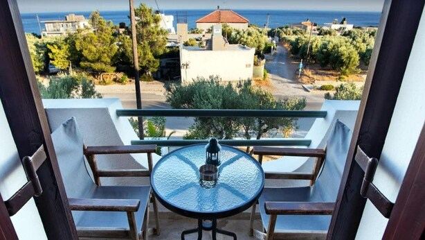 A balcony with a glass table and two chairs, overlooking a coastal area with buildings and trees. - New Aurora By Checkin (Ierapetra)