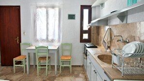 A compact kitchen with a stone floor, a dining table with chairs, a sink with dish rack, and a window with curtains. - New Aurora By Checkin (Ierapetra)