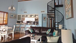 Ein geräumiges Wohnzimmer mit einer braunen Ledersofa, einem Essbereich mit weißen Stühlen, einer Küche im Hintergrund und einer schwarzen Wendeltreppe.