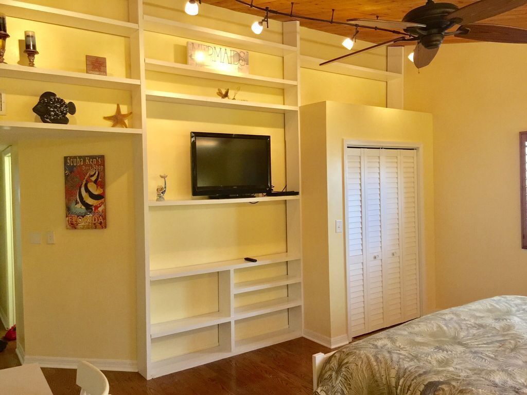 Un dormitorio con cama, televisor en un estante blanco, armario y ventilador de techo.