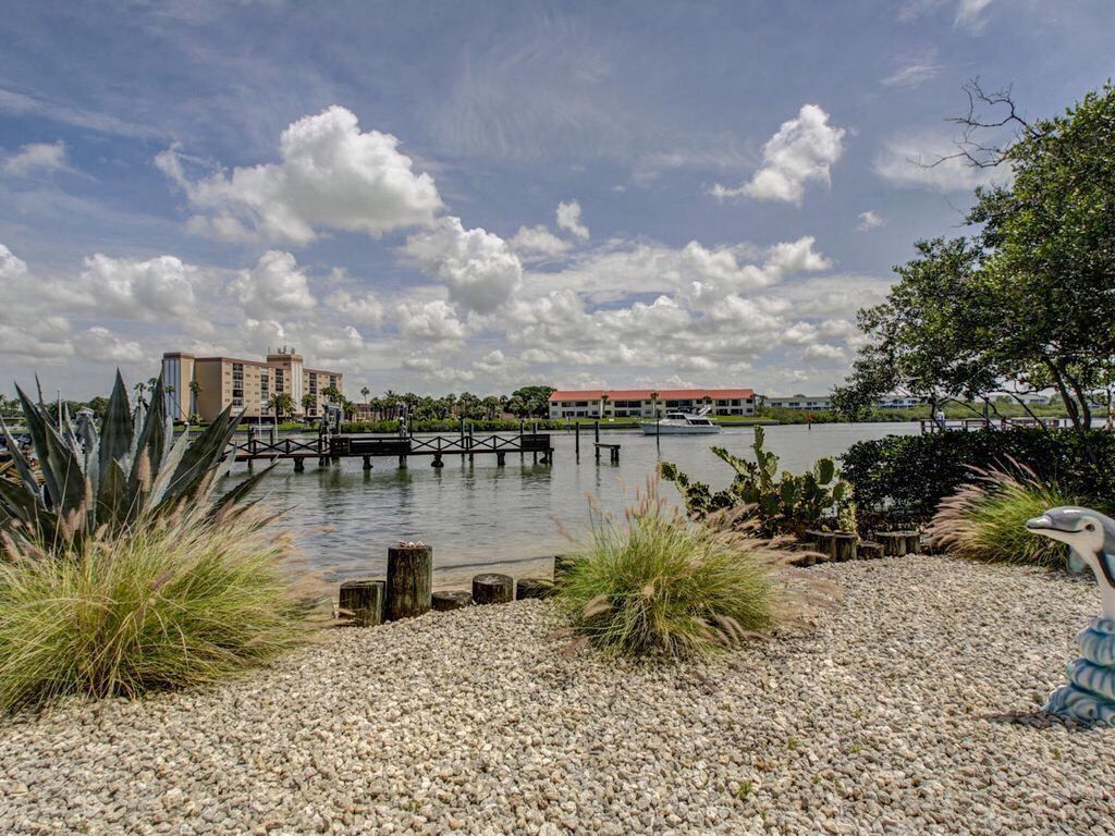 A lakeside view with a dock, pebbled beach, and buildings in the background.