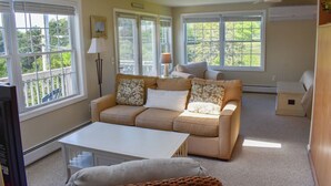 Una sala de estar acogedora con un sofá color beige, una mesa de centro blanca y un televisor. Hay ventanas grandes con persianas, un ventilador de techo y una pequeña mesa con lámpara.