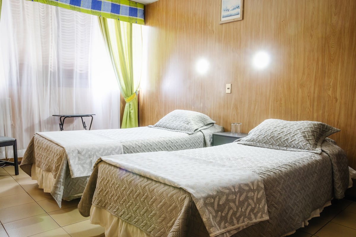A room with two beds, each with a patterned bedspread, a small table, and a chair.