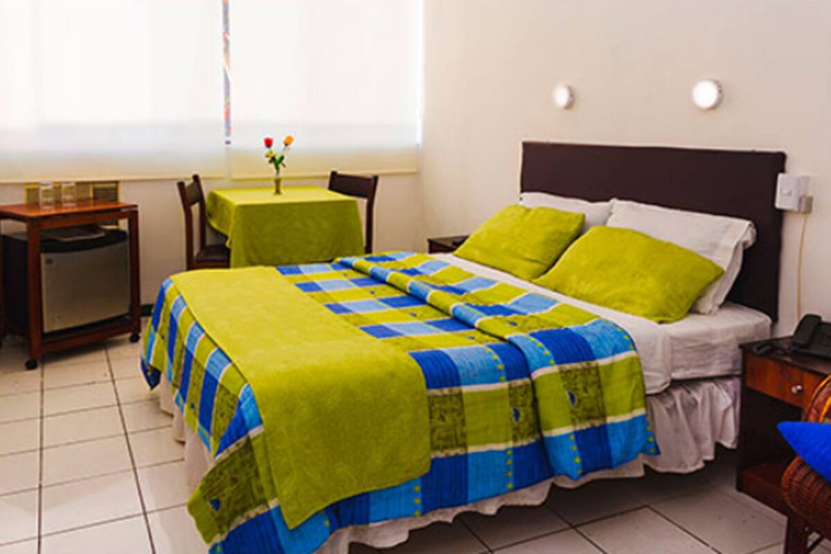 A hotel room with a bed, a small table with a tablecloth, a chair, and a bedside table with a lamp.