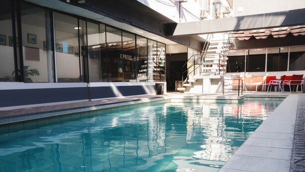 A swimming pool with clear water, adjacent to a building with large windows and a staircase leading to an outdoor seating area with red chairs. - Hotel Concorde (Arica)