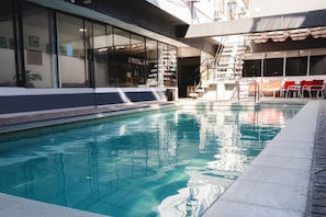 A swimming pool with clear water, adjacent to a building with large windows and a staircase leading to an outdoor seating area with red chairs. - Hotel Concorde (Arica)