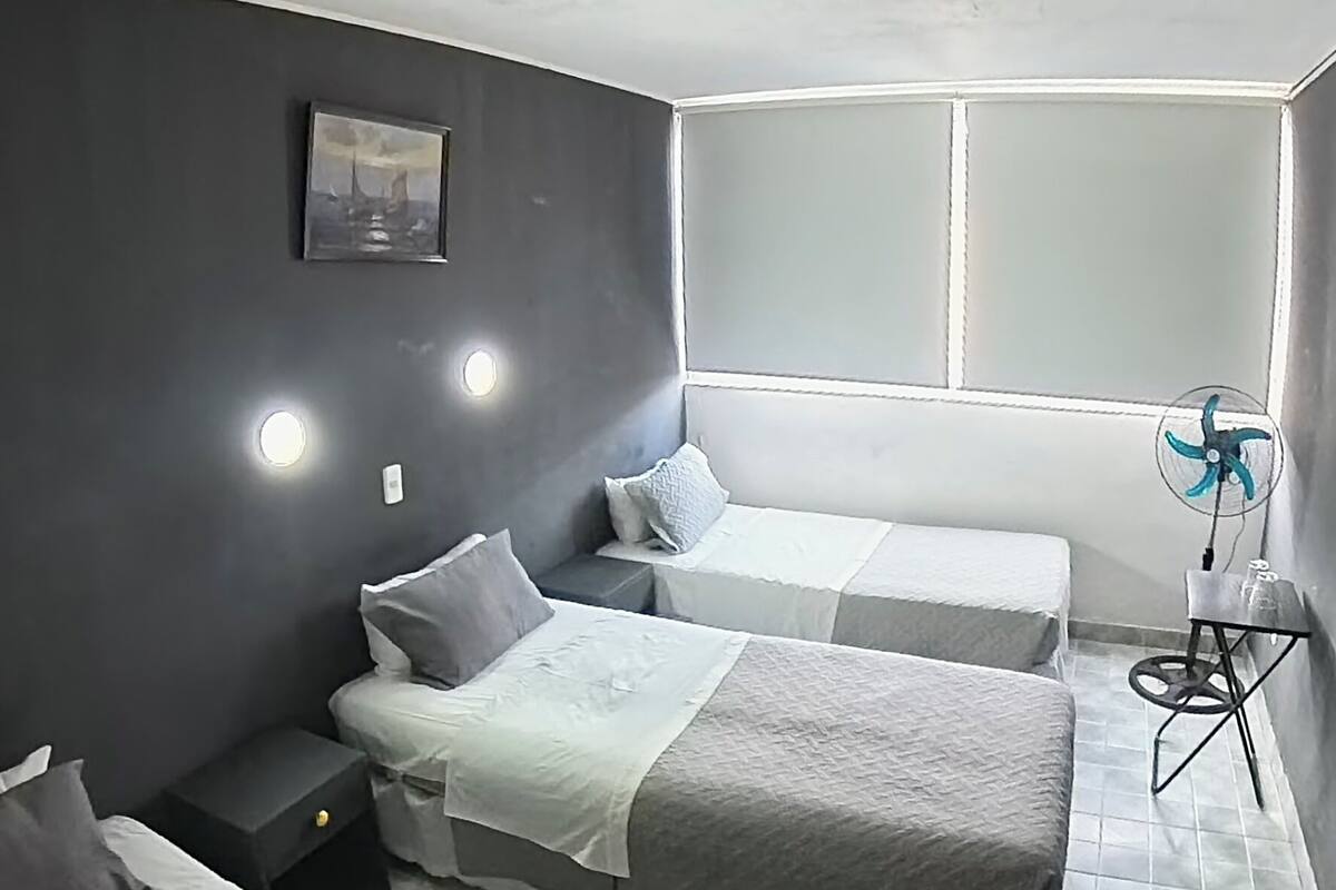 A bedroom with two beds, a small table, a fan, and a picture on the wall.