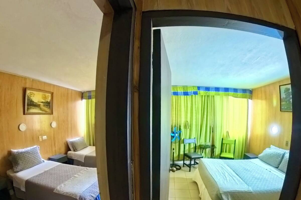 A room with two beds, a wooden floor, and a view through an archway to another room with a bed and a fan.