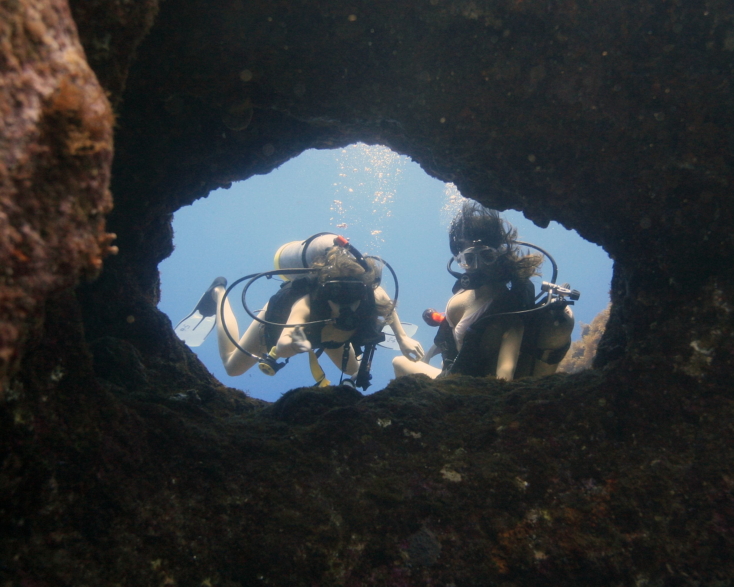 Two divers inside a cave with visible bubbles and a clear blue sky above.