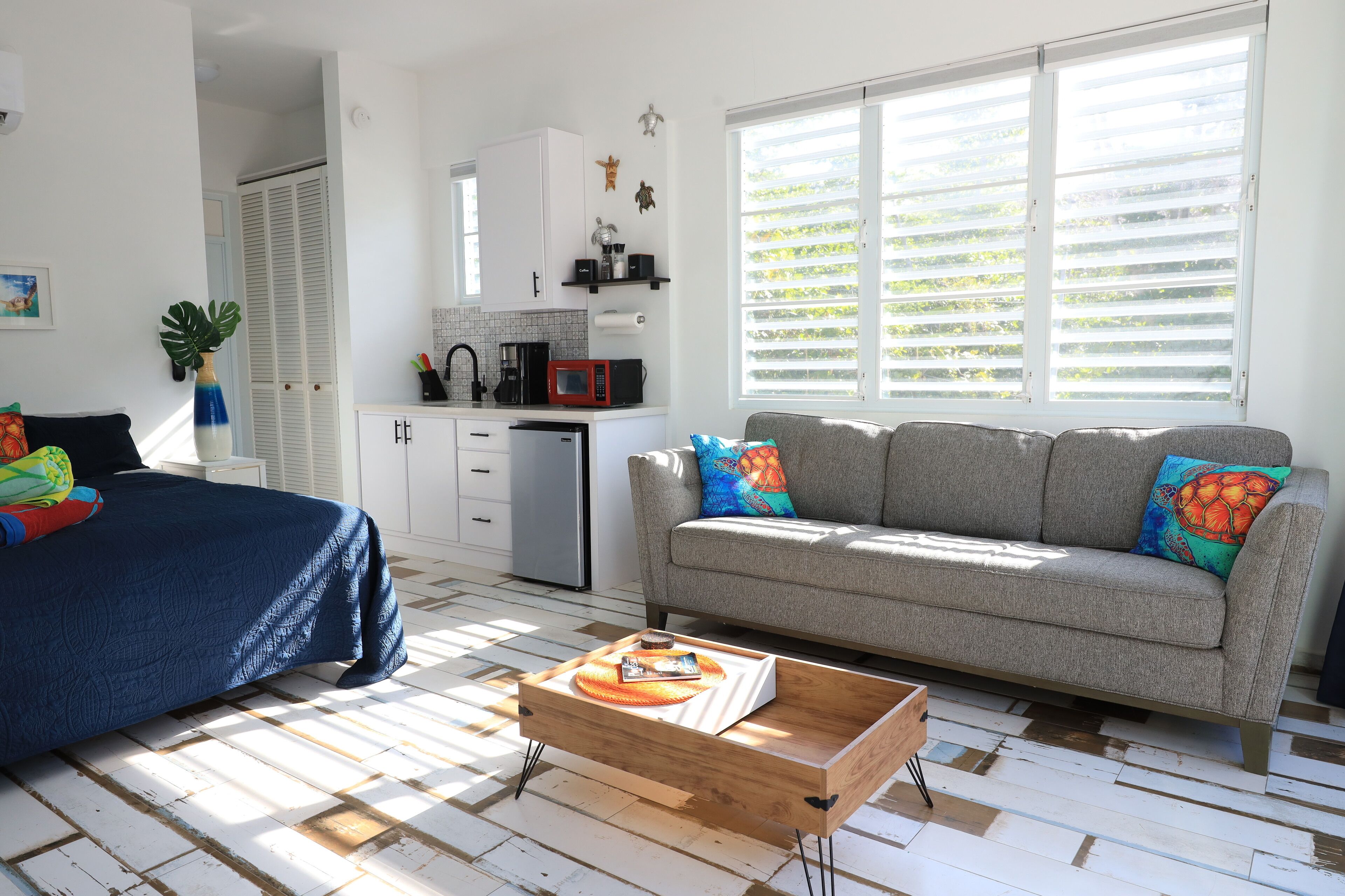 A room with a bed, a sofa, a wooden coffee table, a kitchenette, and a window with blinds.