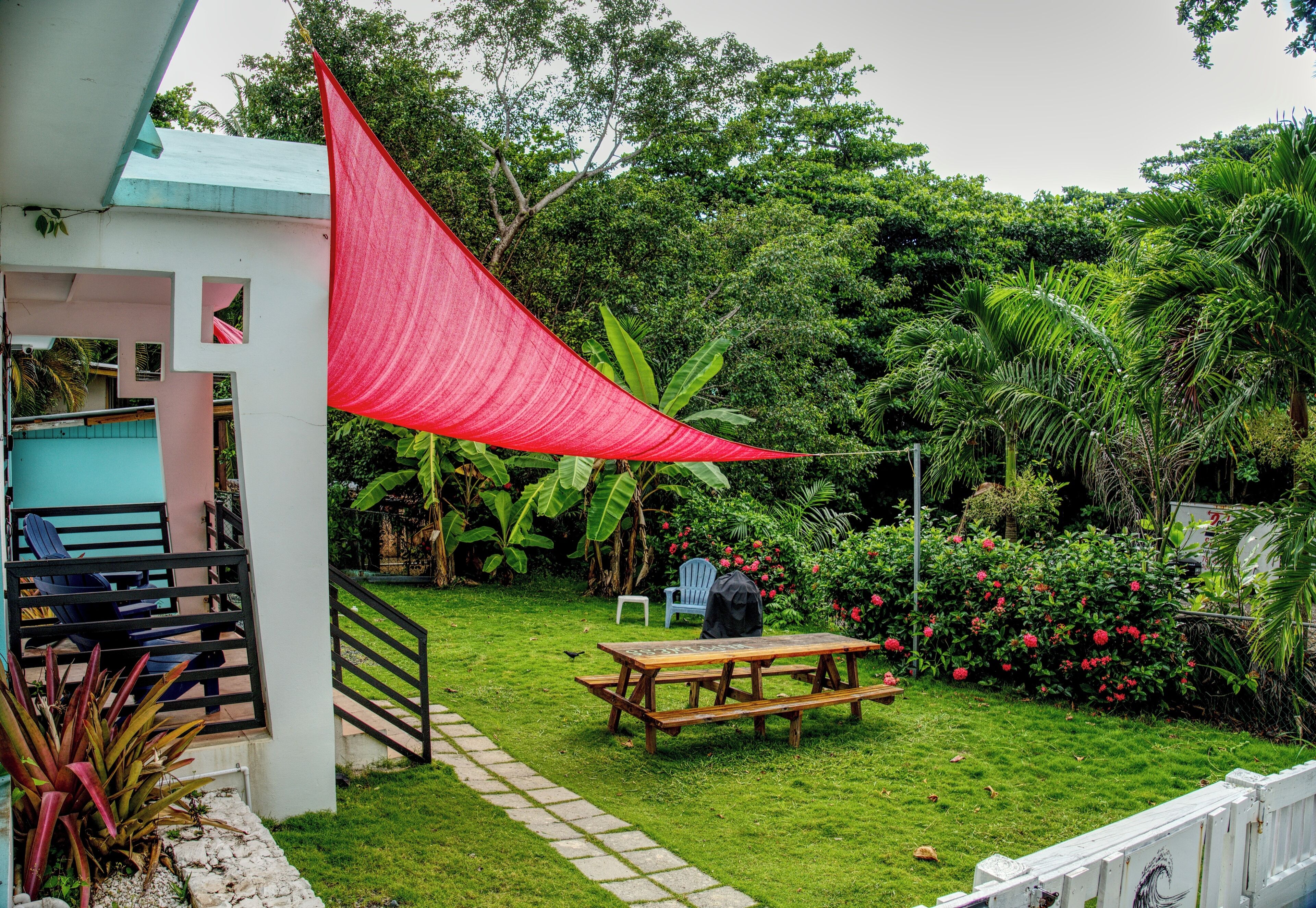A patio with a wooden table and chairs, surrounded by lush greenery and a red shade sail.
