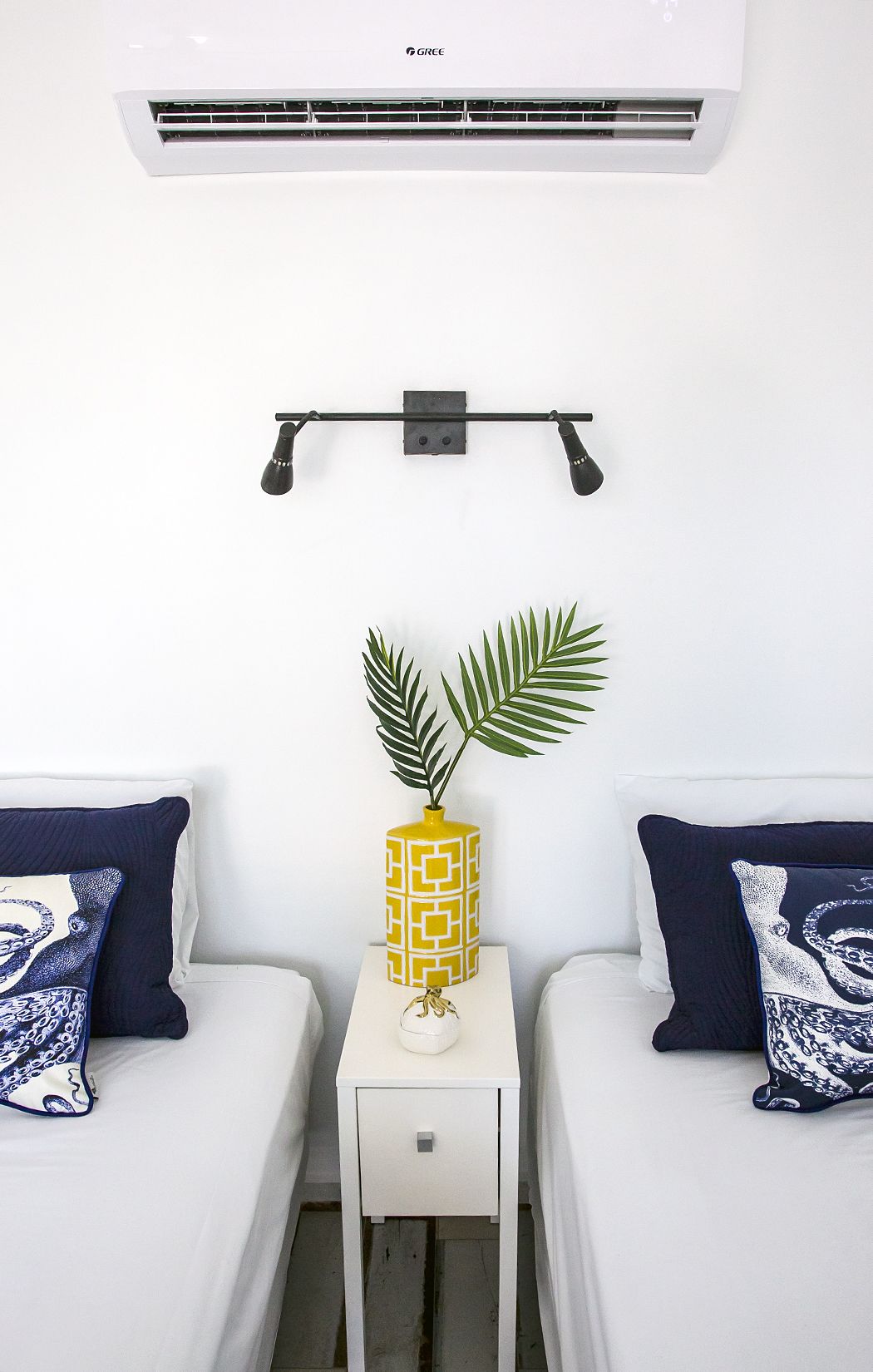 A hotel room with two beds, a small white nightstand, a wall-mounted air conditioner, and a shelf with a decorative vase and plant.