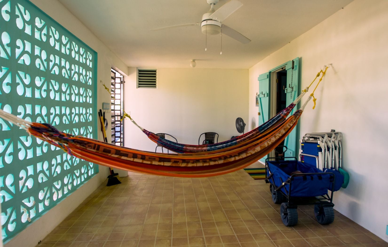 A hammock in a room with tiled flooring, a ceiling fan, a window with blinds, and a blue cart with wheels.