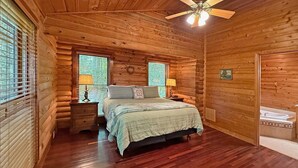 Une chambre à coucher située dans un chalet en rondins, avec un lit, deux tables de chevet, un ventilateur de plafond et une fenêtre avec des stores.