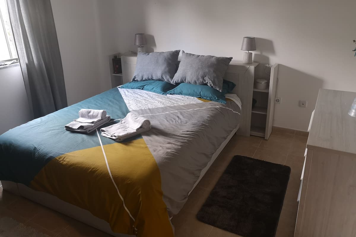 Una cama bien hecha con colcha azul y amarilla, almohadas grises y una toalla doblada al lado. Hay dos mesitas de noche con lámparas, un estante pequeño y una alfombra oscura en el suelo.