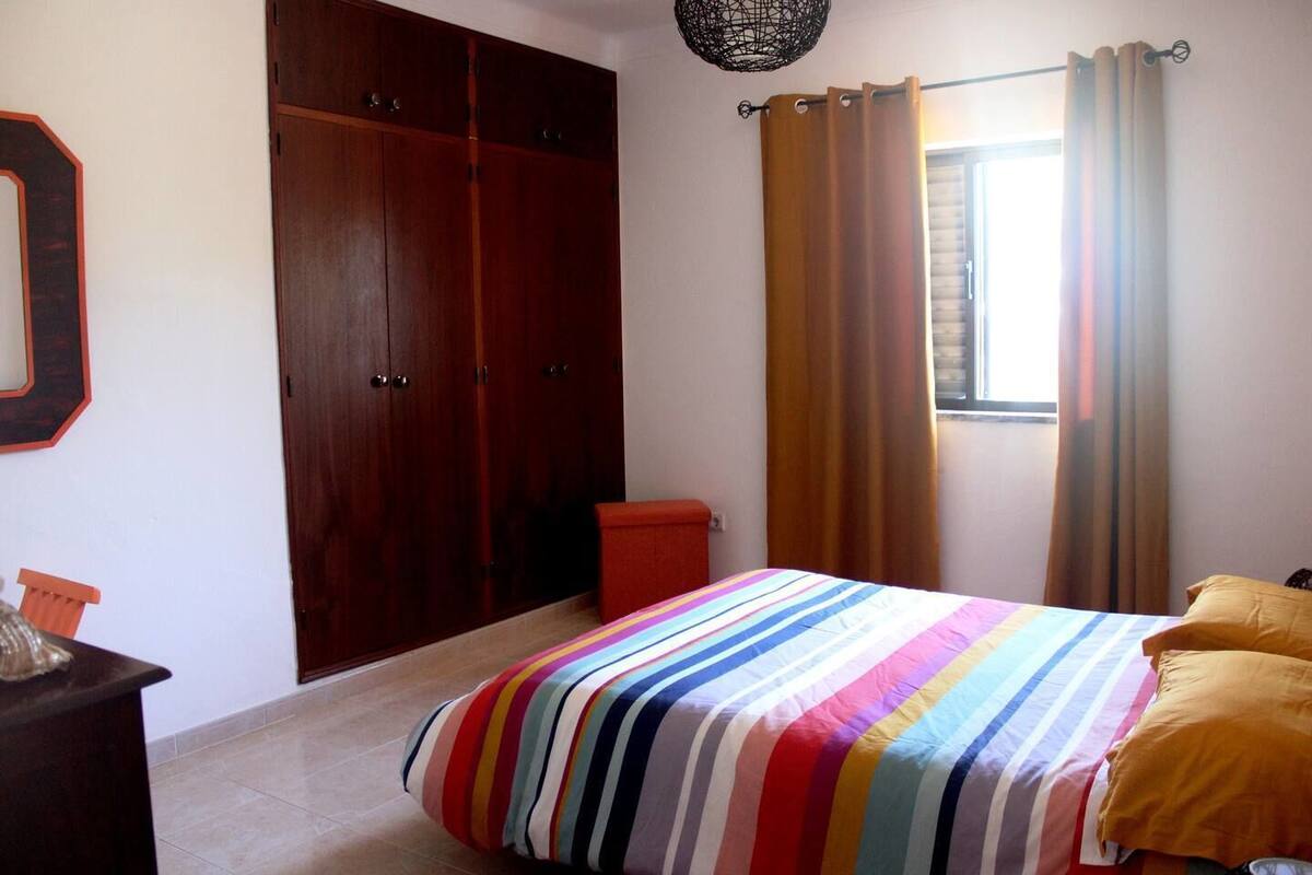 Un dormitorio con edredón a rayas, un armario de madera, un taburete rojo y una ventana con cortinas.