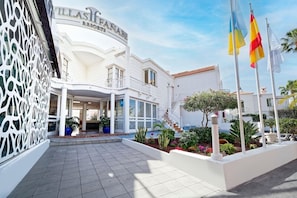 A white building with a sign reading 'VILLAS FANABES RESORTS.' There is a covered entrance, a staircase leading to the entrance, and a landscaped area with plants and a flagpole with flags. - Bluesea Bahia Fanabe and Villas (Adeje)