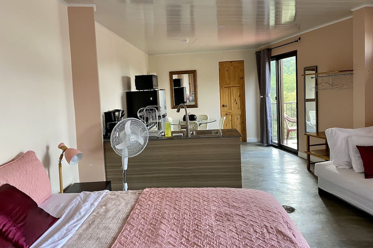 A bedroom with a bed, bedside table, and a fan. There is a dining area with a table and chairs, and a view of the outdoors through a door.