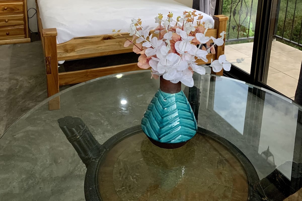 A bedroom with a bed, a nightstand, a vase with flowers, and a view of greenery through a window.
