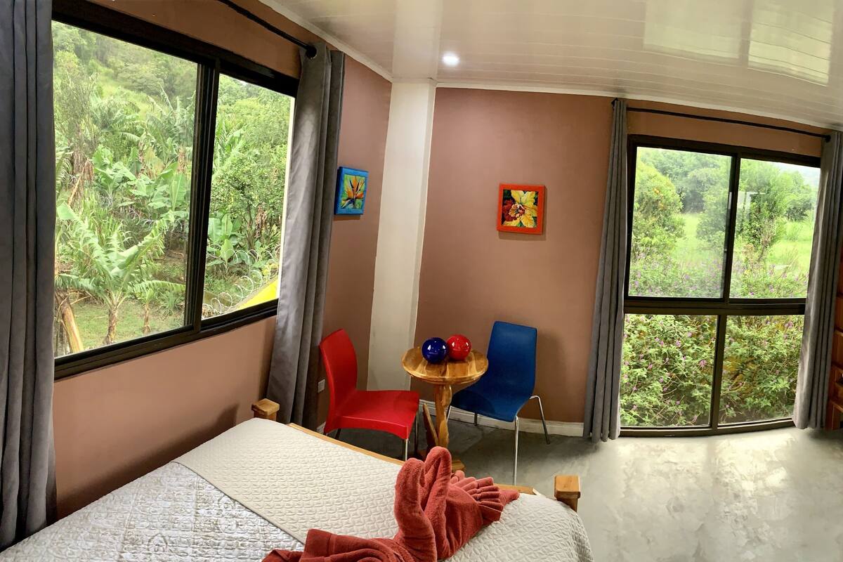 A room with a bunk bed, a single bed, a small table with two red and one blue ball, two chairs, and a large window with a view of greenery.