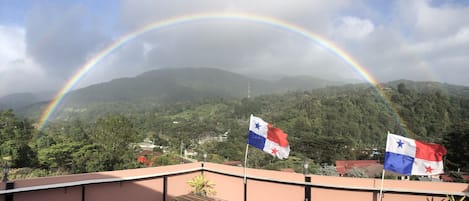 A rooftop terrace with two flags, a wooden table, and a view of a mountain range.