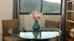 A dining area with a glass table, two chairs, a vase with flowers, and a view of a forest through a large window. - Buena Vista Boquete (Boquete)