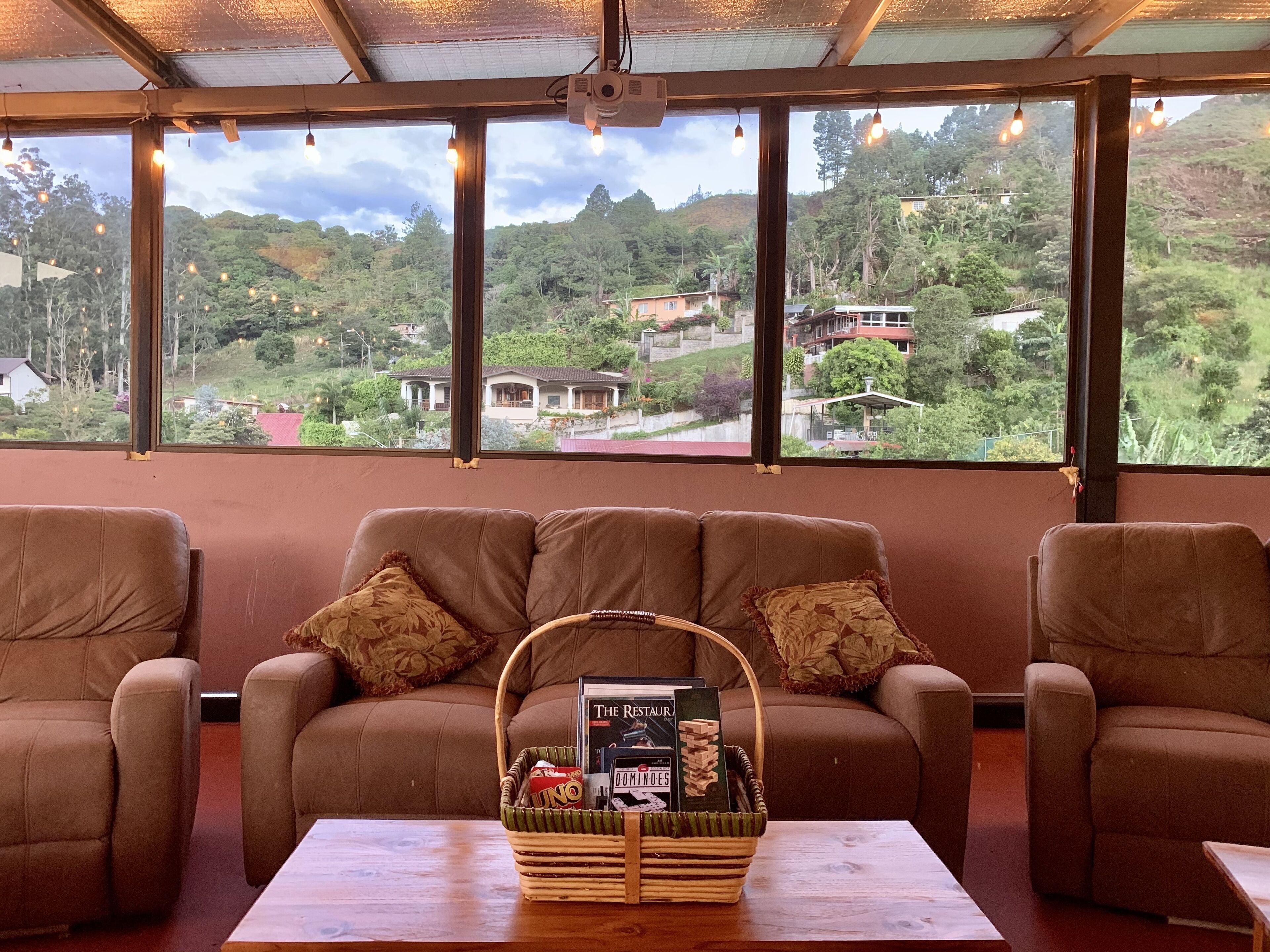 Un coin salon accueillant avec un canapé et des fauteuils, une table basse en bois et un panier rempli de livres et de collations. De grandes fenêtres offrent une vue sur un paysage verdoyant et luxuriant, avec des maisons.