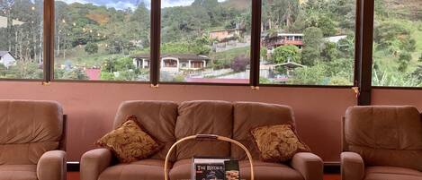 A cozy seating area with a sofa and armchairs, a wooden coffee table, and a basket with books and snacks. Large windows offer a view of a lush, green landscape with houses.