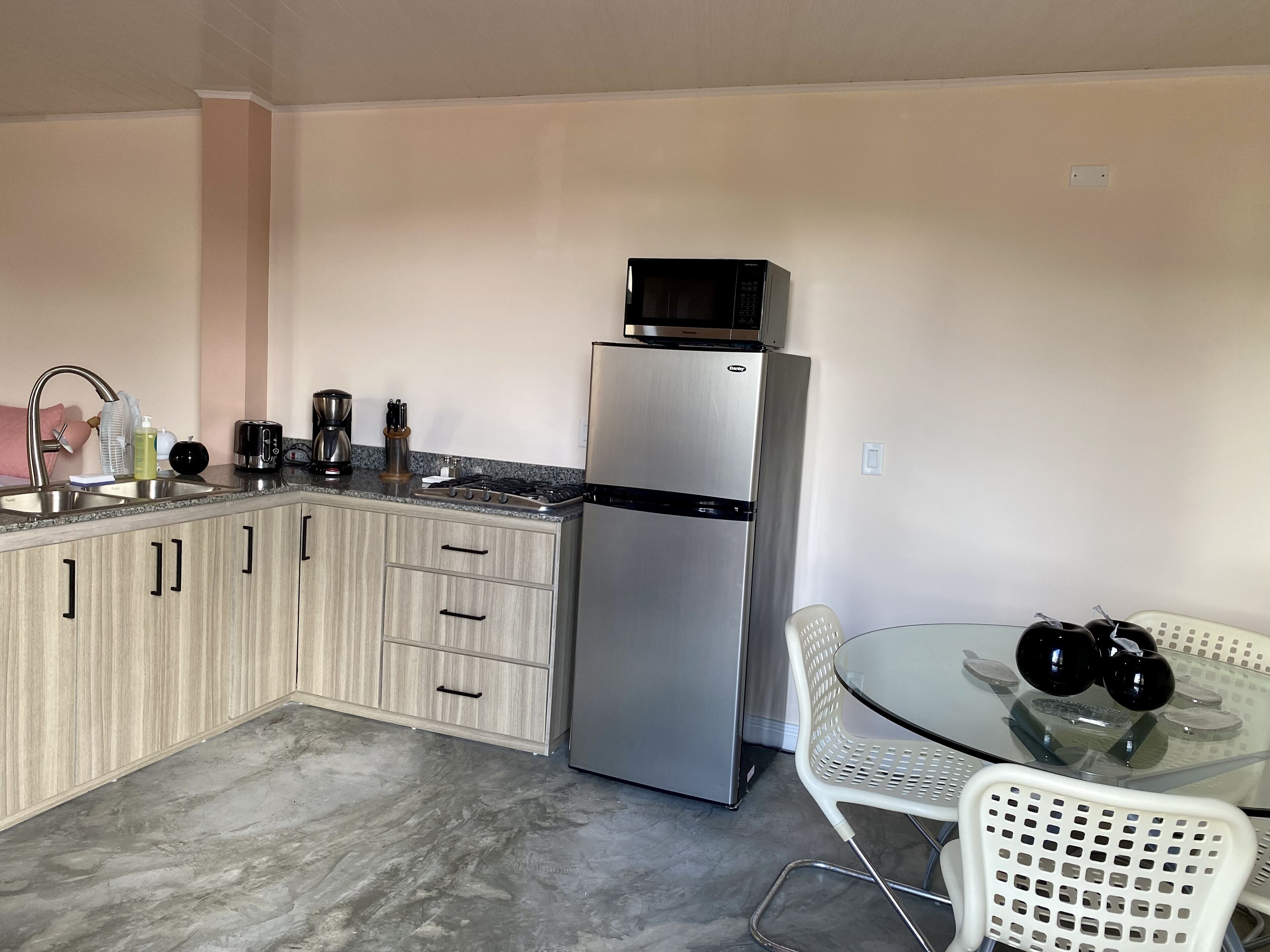 A compact kitchen with a stainless steel refrigerator, a microwave, a sink, and a small dining area with a glass table and white chairs.