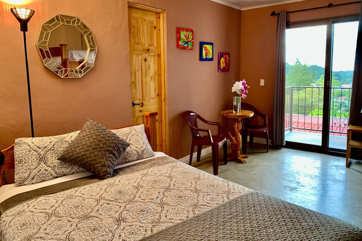A bedroom with a bed, a round table with chairs, a wooden door, and a balcony with a railing.